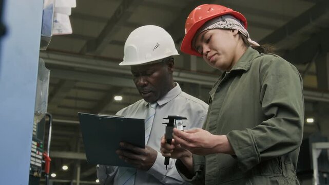 Low angle shot of diverse male and female supervisor and production engineer scaling metal detail with vernier calliper and writing results into quality report in manufacturing plant