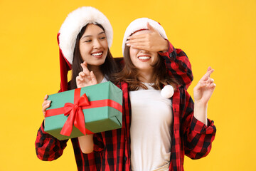 Young lesbian woman surprising her wife with Christmas gift on yellow background