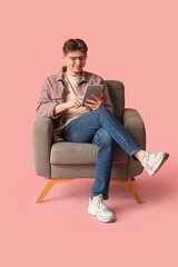 Handsome young man with modern tablet computer sitting in armchair on pink background