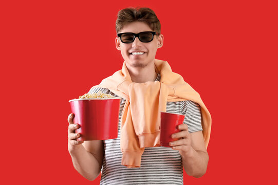 Handsome young man in 3D glasses holding bucket with popcorn and plastic cup of cola on red background
