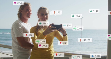Obraz premium Social media notifications image over couple taking selfie by sea