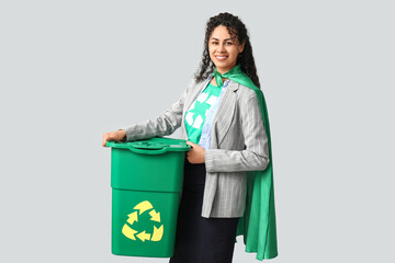 Beautiful young African-American businesswoman in t-shirt with recycling symbol and superhero cape holding trash bin on grey background © Pixel-Shot