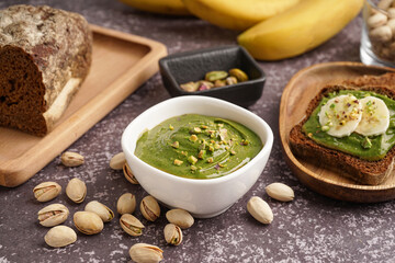 Bowl of tasty pistachio paste and bread with banana slices on grey grunge background