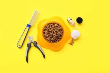 Bowl with dry food and pet care tools on yellow background