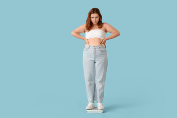 Overweight young sad woman with scales on blue background. Weight gain concept