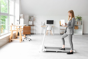 Young businesswoman with laptop training on treadmill in office