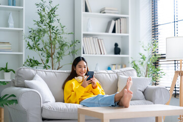 Happy young asian woman  wearing headset and relaxing on comfortable couch at home, texting messaging on smartphone, smiling girl use cellphone chatting, online shopping at home, video call