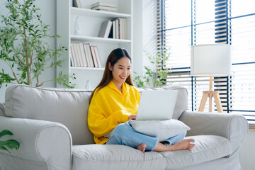 Happy young Asian woman using laptop while seated on couch at home