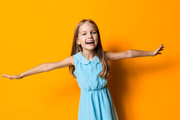 Joyful little girl in a blue dress with arms wide open on a vibrant orange background, celebrating...