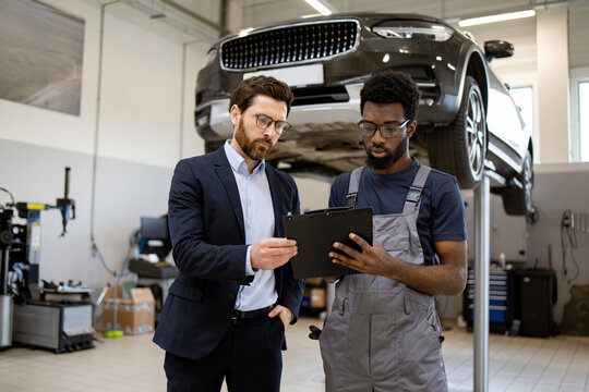 Mechanic in gray overalls showing tablet to businessman in suit inside car repair shop. Discussing vehicle maintenance details. Car on lift in background. Professional cooperation, automotive industry