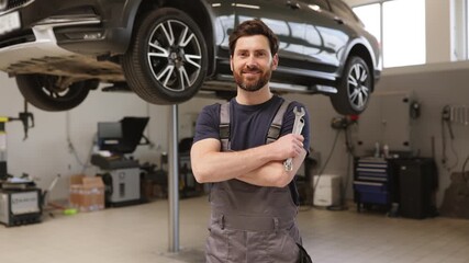 Smiling mechanic holding wrench in modern auto repair shop with car on lift. Confident professional in uniform ready for vehicle maintenance and repair services in clean workshop environment.
