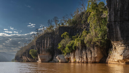 A calm red-brown African river. Green vegetation grows on the steep limestone coastal cliffs. Clouds in the blue sky. Glare on the water. Morning haze in the distance. Madagascar. Manambolo river.