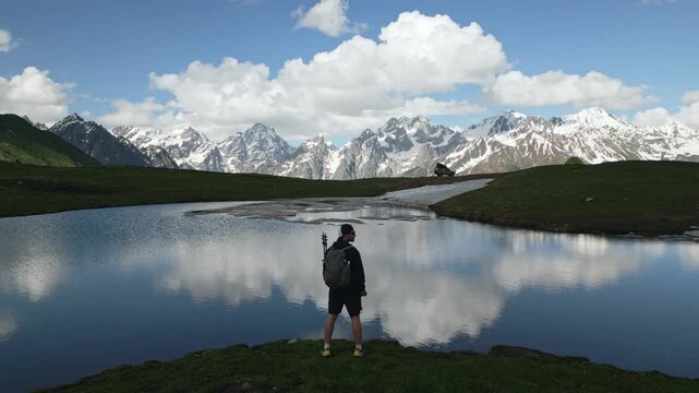 Drone view of hiker with backpack at Koruldi lakes. Stunning mountain landscape. Man explores nature. Adventure, hiking. Reflective water, snow-capped peaks, cloudy sky. Svaneti region, Georgia.
