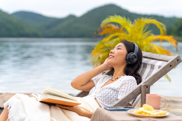 Fototapeta premium Asian woman sitting on lake house balcony reading a book and listening to the music on headphones with mobile app. Attractive girl enjoy outdoor lifestyle travel nature on summer holiday vacation.
