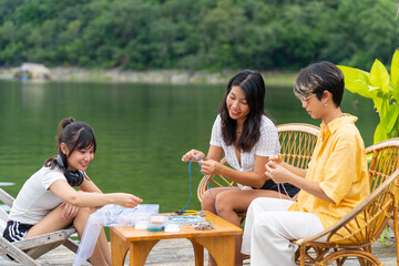 Young Asian woman enjoy and fun outdoor lifestyle travel nature forest mountain on summer holiday vacation. Happy girl friends relaxing with hobbies stringing beads together on lake house balcony.