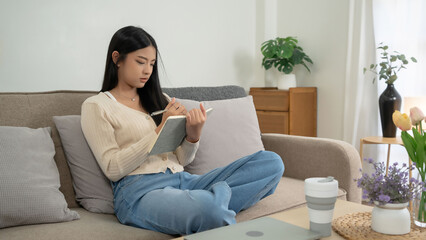 Fototapeta premium Young asian women reading a book to researching educational and taking notes while sitting on comfortable couch to learning self-study and spending time to relaxation with enjoying lifestyle at home