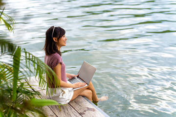 Happy Asian woman using laptop computer remote working online business on on lake house balcony. Attractive girl relax and enjoy outdoor lifestyle travel forest nature on summer holiday vacation.