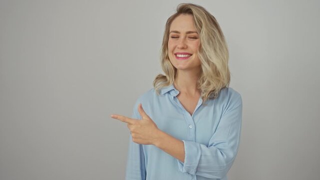 Cheerful young blonde woman wearing shirt, naturally expressing happy emotion with a smug smile while pointing up with a finger at the side. stands isolated against a plain white background.