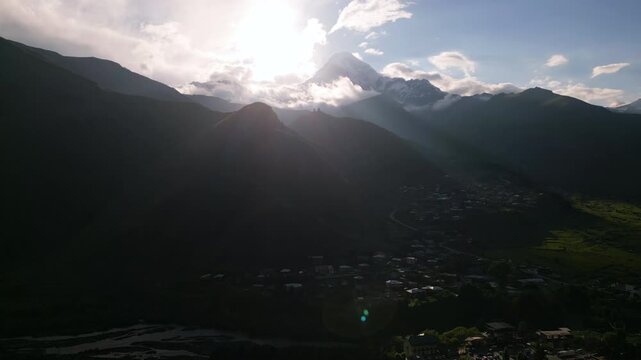 Aerial view of Kazbegi peak in Caucasus mountains with village in shadow and sunlight. Clouds and snowy summit. Drone footage captures scenic mountain landscape.