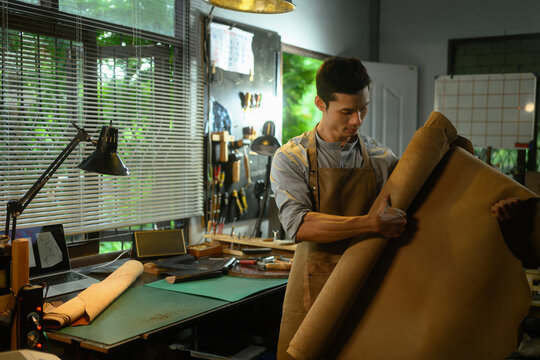 Skilled leather craftsman standing amidst various tools and examining a large roll of leather in his workshop