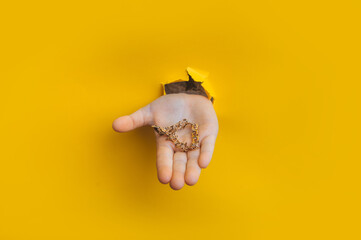 A golden bracelet in a child's hand protrudes through a torn hole in yellow paper. Concept of precious jewelry.