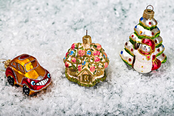 Three colorful and festive Christmas ornaments, including car, decorated house and snowman with Christmas tree, placed on bed of artificial snow