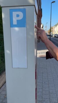 Vertical. Woman is paying for car parking on the city street, pay and display ticket machine or parking meter; side view, handheld shot, Panevezys city downtown, Lithuania, Europe.