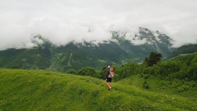 Solo traveler with backpack hikes through alpine meadows, enjoys nature. Explores scenic views, green hills, cloudy mountains. Enjoy adventure, freedom in wild alpine landscape, summer hiking trail.