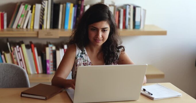 Focused positive young adult Indian student woman working on business project at computer in library, typing on laptop, thinking on online communication, looking away, sitting at workplace table