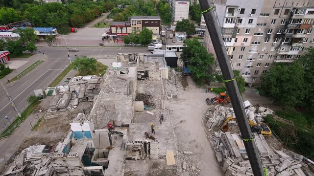 Dismantling war-damaged building with crane lifting concrete slabs. Drone view shows construction workers, demolition of residential structure near urban streets. Rebuilding area after war damage.