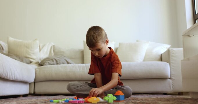 Focused cute little kid boy playing creative construction game at home, stacking toy building blocks on warm carpeted floor, enjoying childhood, smart activity, playtime. Low angle view