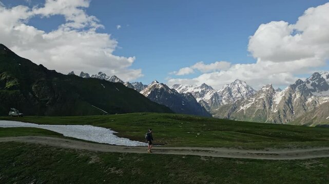 Hiker walks alpine meadows in high mountains. Snow patches on green hills. Majestic peaks under blue sky with clouds. Outdoor adventure, nature exploration. Scenic landscape, summer hiking route.