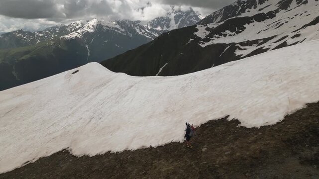 Aerial view of man with backpack descending snowy slopes in Svaneti mountains. Alpinist hiking, exploring rugged terrain. Outdoor adventure, active leisure, mountain climbing in remote area. Drone