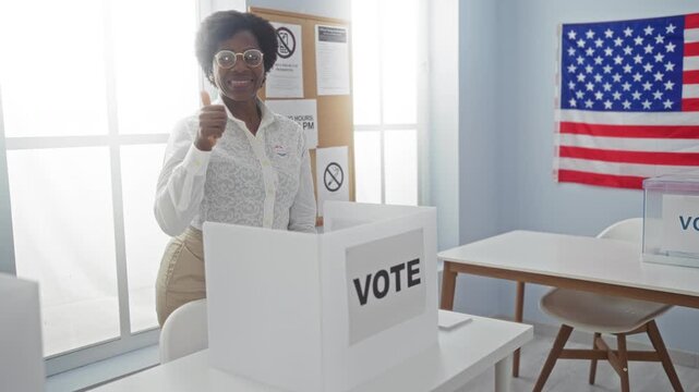 African american woman gesturing thumbs up at a voting booth with a usa flag in the background inside an electoral college room.