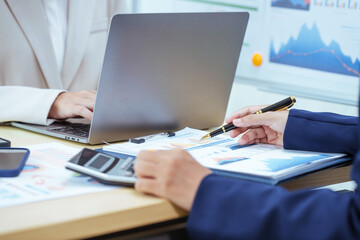 Two young Asian businesswomen sit at a table, engaged in a meeting.examine graphs and discuss capital investment strategies, showcasing their expertise and collaborative approach to business growth.