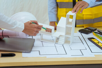 Two young women work at a desk, meticulously designing and drawing plans for building a house. As...