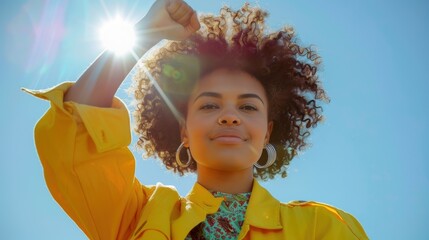 Confident woman with curly hair and yellow jacket raising fist in sunlight against blue sky backdrop, demonstrating strength and empowerment.