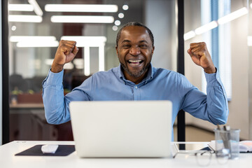 Excited office worker celebrating success at desk in modern workplace. Businessman raising fists in triumph while working on laptop. Achievement and positive emotion in professional environment