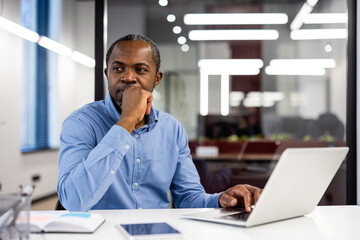 Pensive office worker in blue shirt thinking at desk with open laptop and documents. Modern office environment with bright lights and large windows. Concept of contemplation, problem-solving