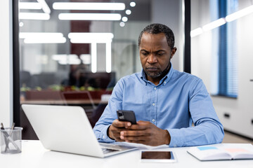 Businessman in blue shirt sitting at office desk using smartphone while working on laptop. Modern office environment with professional focus and productivity