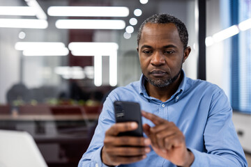 Office worker using mobile phone in modern workplace. Concentrated businessman checking email on smartphone during work hours. Professional in business attire interacting with digital device.
