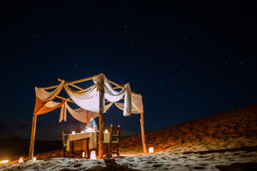 A vibrant desert camp with traditional Moroccan tents, adorned with colorful rugs and cushions. Palm trees and drums create a lively atmosphere, set against a backdrop of rolling hills and a clear blu © LAHCENFOTOS