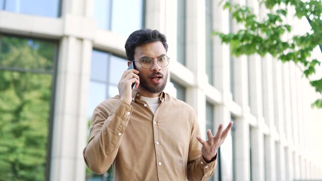Angry excited businessman arguing talking on a mobile phone while standing on street near an business office building. Annoyed manager quarrel on the smartphone, describing a problem to be solved