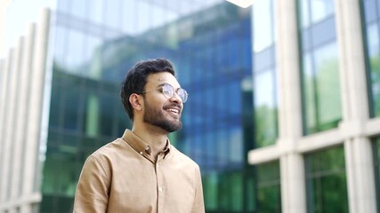 Confident businessman walks down street near an office building. Bearded entrepreneur in glasses walking in central business district of modern city. Handsome man worker goes to work. Close up 