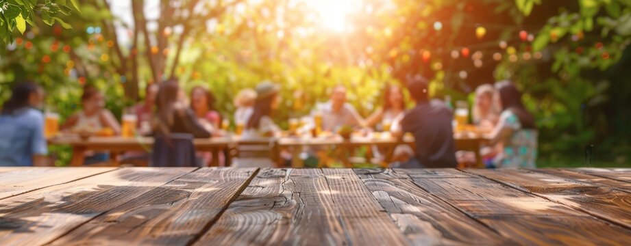 Empty wooden table and blurred view of a group of people having BBQ barbecue outdoors. Wood desk in front of a natural garden background. Summer lifestyle concept.