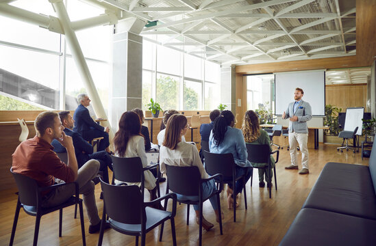 Young berded man speaking on a business training standing in office during a conference or seminar for a group of diverse people company employees sitting on chairs in meeting room.