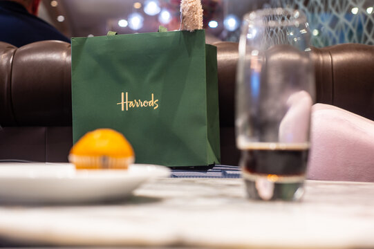 London, UK. July 23 2023. A green Harrods shopping bag stands out in a cozy caf&eacute; setting with a muffin and drink in the foreground.
