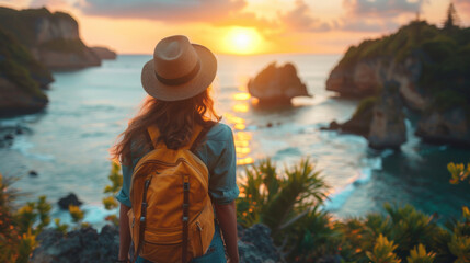Fototapeta premium Back view of a traveler with a backpack and hat admiring a beautiful sunset over the ocean, surrounded by lush greenery.
