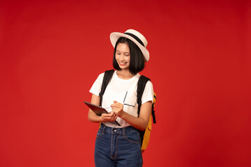 Asian female student wearing white hat presenter of university and tutoring institute, carrying bag, using laptop with pen to study online and taking photo in studio on red background.