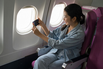 Asian traveler women looking and taking photo of sky view on smartphone through the windows on cabins flight with happiness while sitting near windows on business travel class flight in airplane